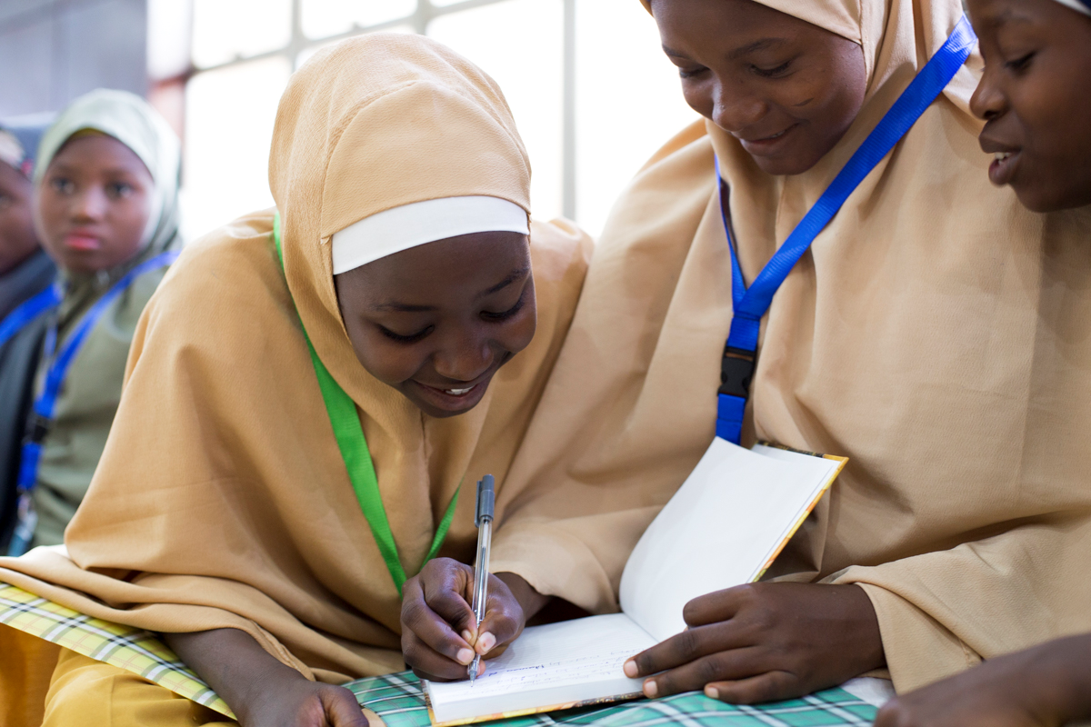 Image of two girls wearing hijabs, one is holding a book and writing while the other, smiling, leans over to see what she is doing