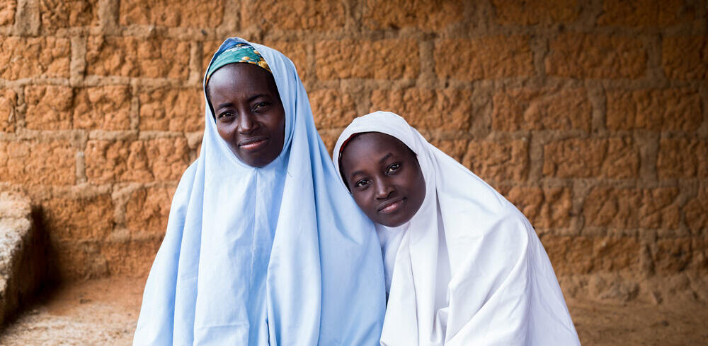 Two women in white hijab smiling, one leaning her head on the shoulder of another